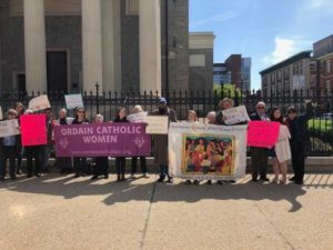 Line of people standing in front of fence by church, with signs saying "Ordain Catholic Women"