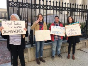 Four adults in front of a black fence, holding signs in support of women's ordination.