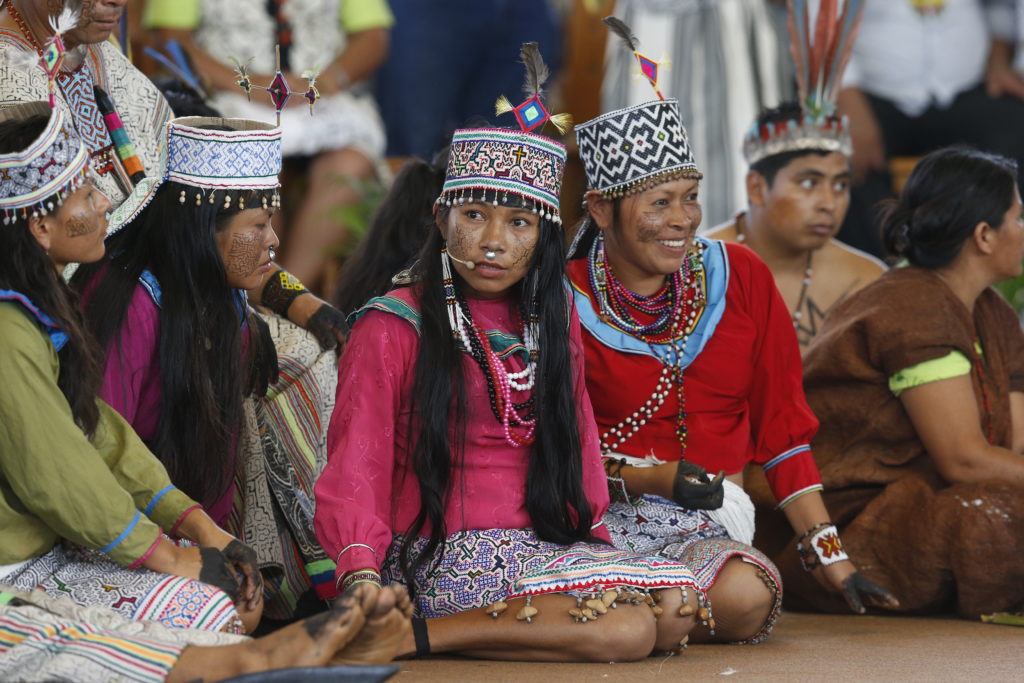 A group of indigenous women from the Amazon sit on the ground, wearing colorful traditional clothing. 