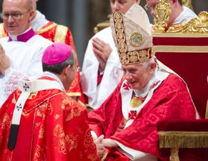 Pope Benedict greets Cardinal William Lori, who is wearing the pink cap of a cardinal. Both men are wearing red liturgical vestments.